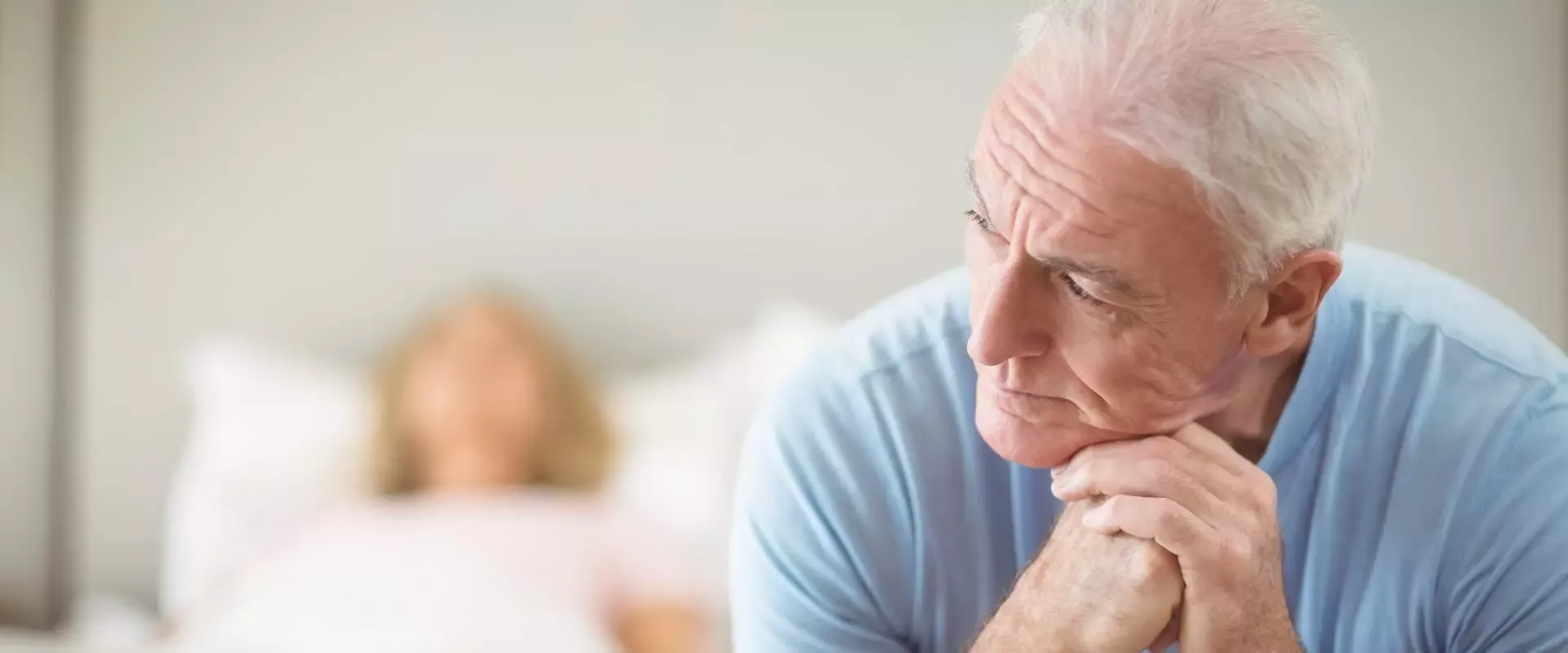 Mann sitzt grübeln am Bettrand, im Hintergrund liegt eine Frau im Bett