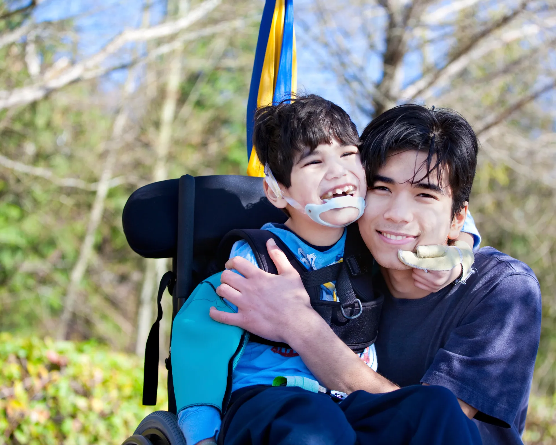 zwei Geschwister, der kleine Bruder sitzt im Rollstuhl, der größere Bruder legt den Arm um ihn