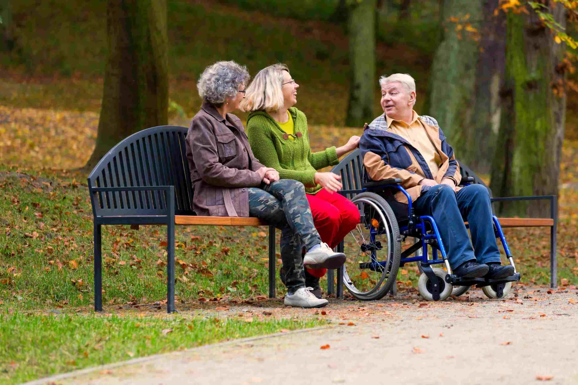 Zwei Frauen sitzen auf einer Park-Bank, daneben sitzt ein Mann im Rollstuhl. Sie unterhalten sich.