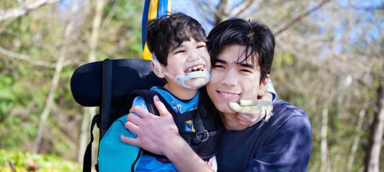 zwei Geschwister, der kleine Bruder sitzt im Rollstuhl, der größere Bruder legt den Arm um ihn