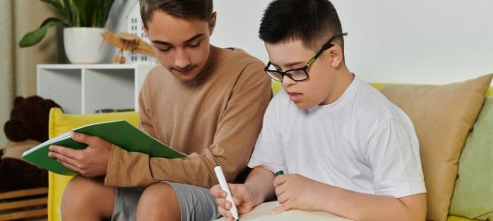 Zwei Brüder sitzen auf der Couch, einer hat das Dwon-Syndrom, der Bruder hilft bei den Hausaufgaben