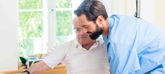 Ältere Mann wird von einem Pfleger aus dem Bett geholfen.