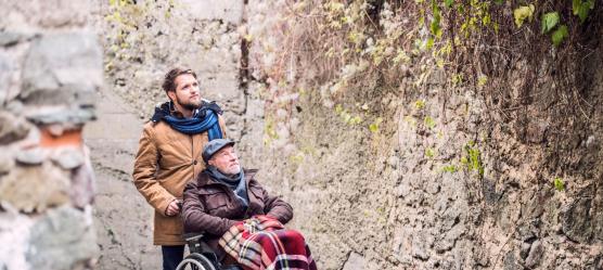Mann schiebt Rentner im Rollstuhl durch eine Straße mit alter Mauer. Beide sind warm angezogen.
