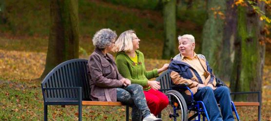 Zwei Frauen sitzen auf einer Park-Bank, daneben sitzt ein Mann im Rollstuhl. Sie unterhalten sich.