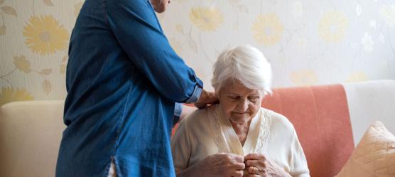 Ältere Frau sitzt auf einem Bett und bekommt Hilfe beim Anziehen.