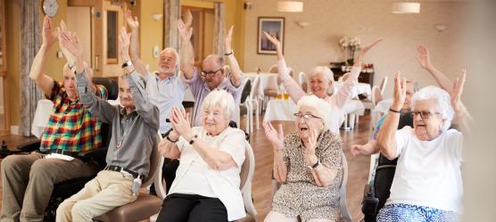 Eine Gruppe in der Tagespflege macht Bewegungsübungen auf einem Stuhl.